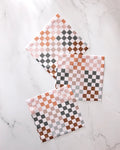 Three GEOMETRY Autumn Checkers square napkins in white, brown, and gray checkered patterns are arranged in a casual, overlapping layout on a white marble surface. This set features absorbent kitchen towels.