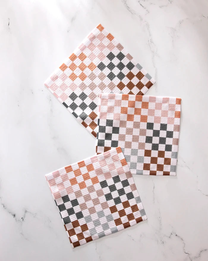 Three GEOMETRY Autumn Checkers square napkins in white, brown, and gray checkered patterns are arranged in a casual, overlapping layout on a white marble surface. This set features absorbent kitchen towels.