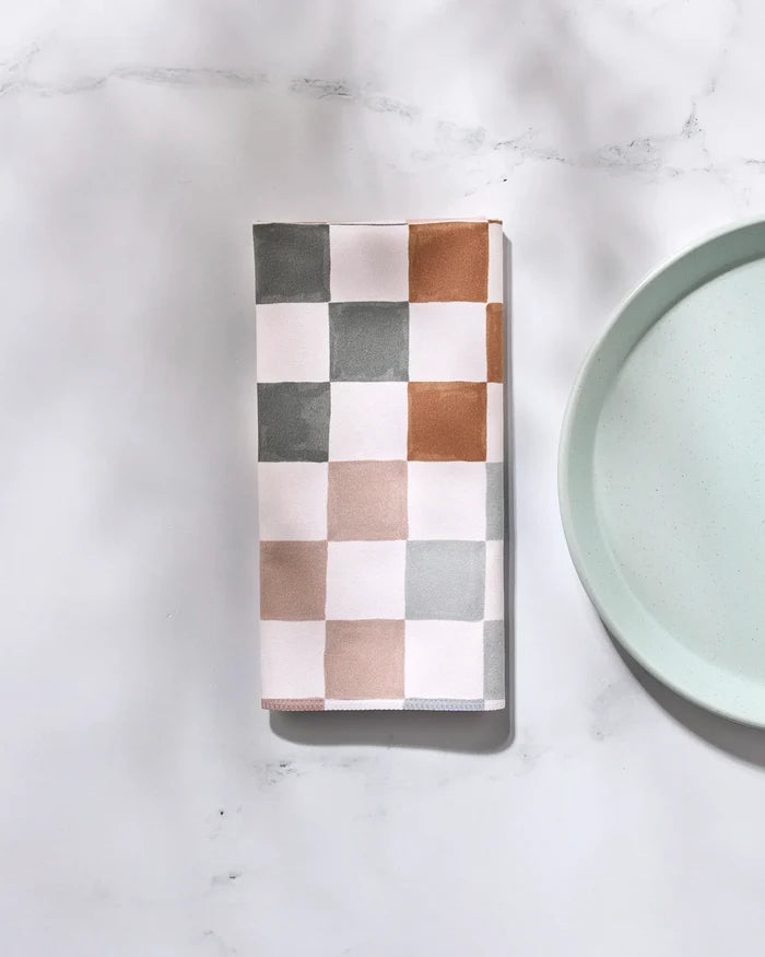 The GEOMETRY Autumn Checkers folded reusable napkin, featuring gray, brown, and beige squares, rests on a white marble surface beside a light blue plate.
