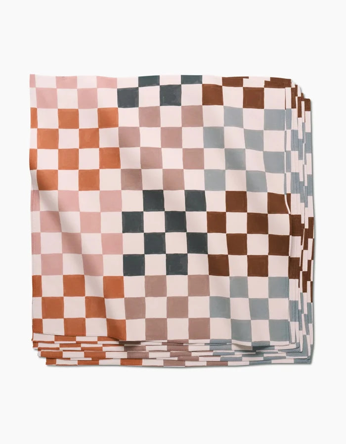 A neatly folded stack of GEOMETRY Autumn Checkers Dinner Napkins, showcasing a checkered pattern in muted pink, brown, gray, and white tones, made from recycled materials and set against a white background.