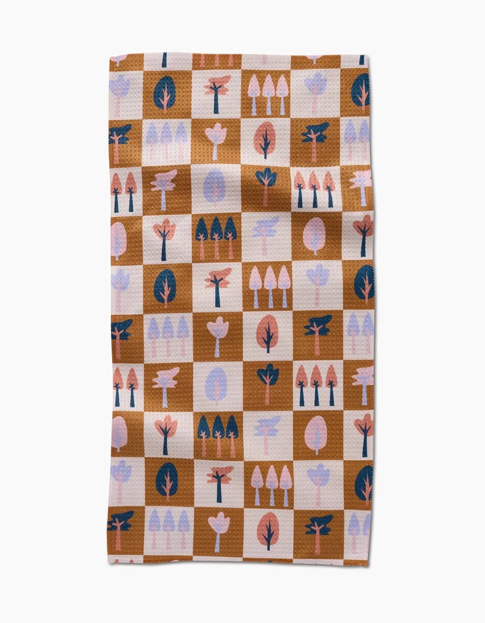 The Autumn Forest kitchen towel by Claire Iglesias is a rectangular recycled towel with a brown and beige checkered pattern, featuring stylized tree illustrations in pink, coral, navy, and light purple on each square.