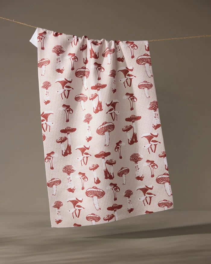 The Geometry Autumn Mushrooms tea towel, made from recycled materials and featuring a red mushroom and animal pattern, hangs on a string against a neutral background.