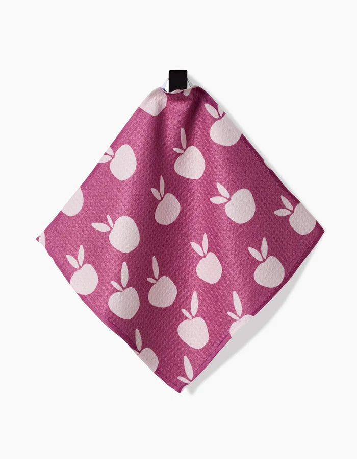 The “Autumn Party” reusable dishcloth by Claire Iglesias, in pink with a white apple and leaf pattern, hangs by a black loop against a white background.