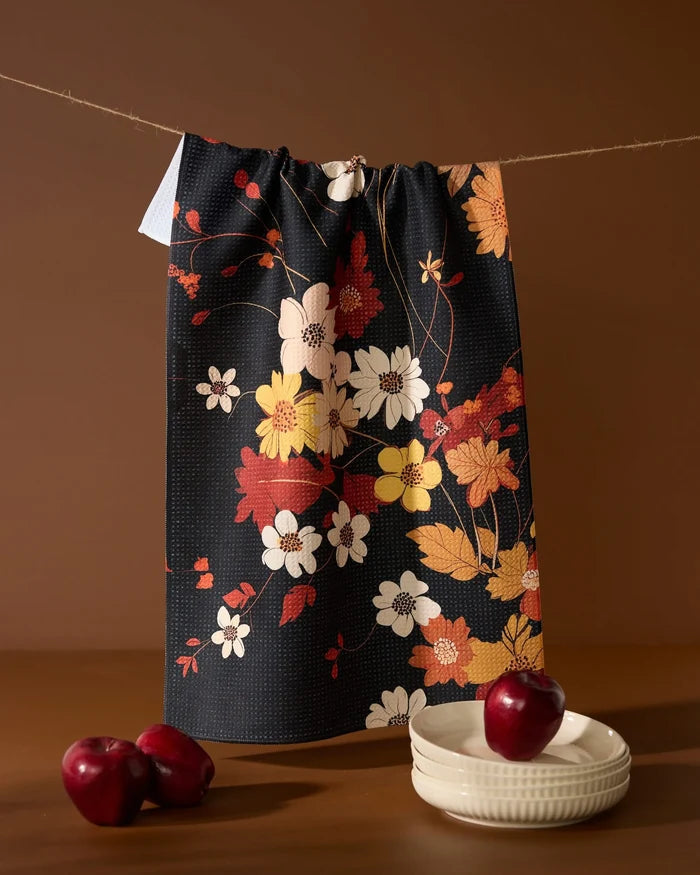 The Geometry Autumn Whispers recycled kitchen towel, featuring a dark floral pattern with red and white flowers, hangs on a string above two white bowls and two red apples set against a brown background.