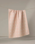 The Aveline tea towel by Amy MacCready, featuring a light pink textured fabric with small white cross patterns, hangs from a clothesline against a neutral background.