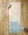The GEOMETRY Balance yoga mat with a light pink to light blue gradient rests on a wooden floor, accompanied by a non-slip yoga towel, water bottle, and two yoga blocks in the mat’s corners.