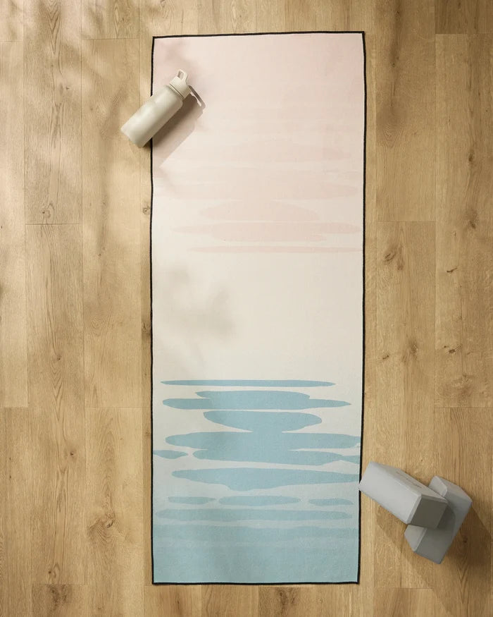 The GEOMETRY Balance yoga mat with a light pink to light blue gradient rests on a wooden floor, accompanied by a non-slip yoga towel, water bottle, and two yoga blocks in the mat’s corners.