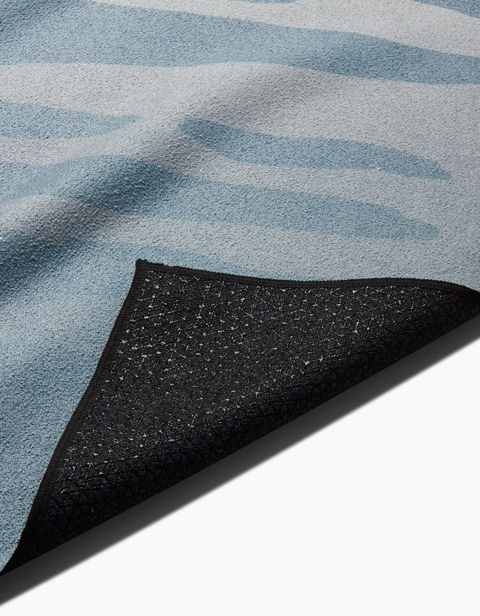A close-up of the GEOMETRY Balance rug shows its blue pattern and a flipped corner, revealing a textured black non-slip backing inspired by slip-resistant yoga towel materials.