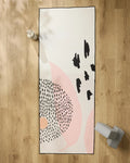 The GEOMETRY Bare rectangular recycled yoga towel, featuring abstract black and pink patterns, is laid on a wooden floor with a white water bottle at the top left and gray weights at the bottom right.