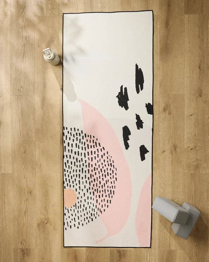 The GEOMETRY Bare rectangular recycled yoga towel, featuring abstract black and pink patterns, is laid on a wooden floor with a white water bottle at the top left and gray weights at the bottom right.