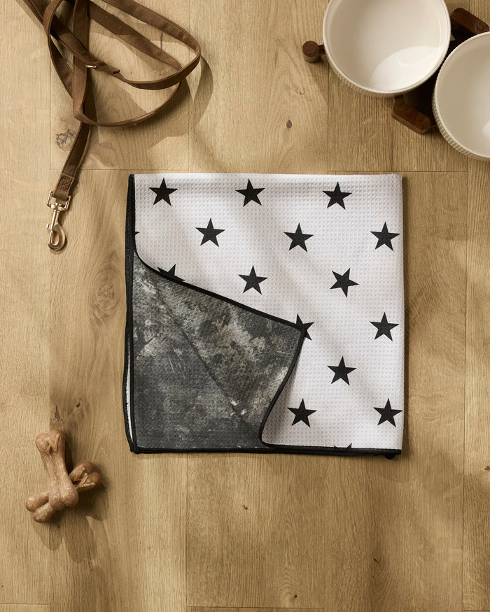 The Geometry Bark white absorbent dog towel with black stars is folded on a wooden floor near two empty pet bowls on a wooden stand, a brown dog leash, and two bone-shaped dog toys.