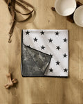 The Geometry Bark white absorbent dog towel with black stars is folded on a wooden floor near two empty pet bowls on a wooden stand, a brown dog leash, and two bone-shaped dog toys.