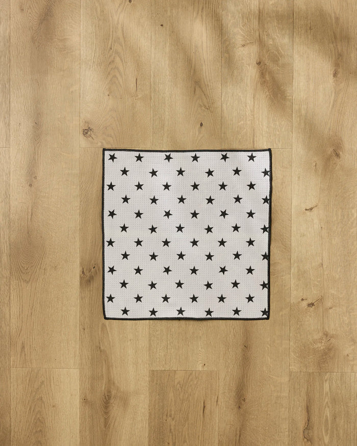 The Geometry Bark towel, ultra-absorbent and eco-friendly, features a white square design with evenly spaced black stars and black borders—perfect for pets. Made from recycled materials and pictured here on a light wood floor.