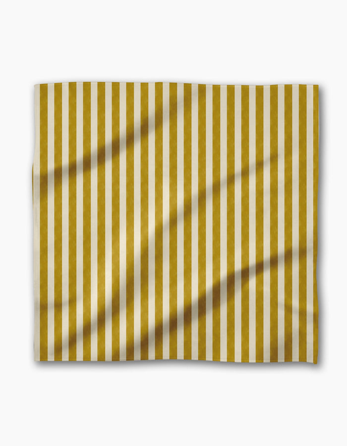 The Geometry Bauhaus Stripe dinner napkin features vertical gold and white stripes on Geosuede fabric, with a slightly wrinkled rectangular shape, displayed against a plain white background.