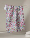 The Bazaar kitchen tea towel by Maayan Alper-Swan features a vibrant pink, green, and blue floral paisley pattern. Made from recycled materials, it brightens kitchens as it hangs near stacked white dishes against a neutral backdrop.