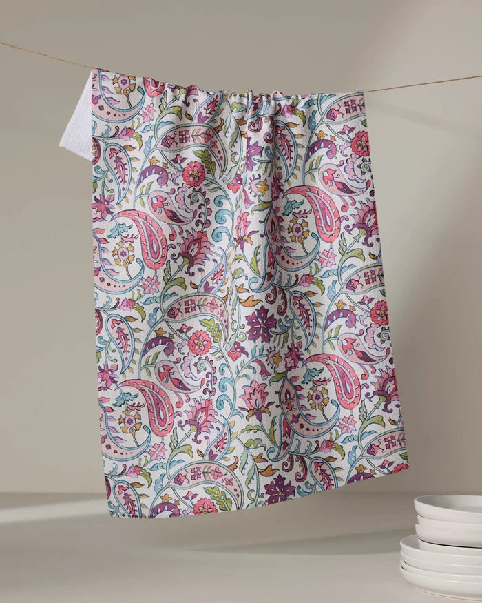 The Bazaar kitchen tea towel by Maayan Alper-Swan features a vibrant pink, green, and blue floral paisley pattern. Made from recycled materials, it brightens kitchens as it hangs near stacked white dishes against a neutral backdrop.