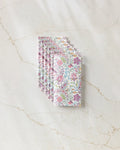 Six folded, eco-friendly Bazaar kitchen towels by Maayan Alper-Swan with a pink, purple, and green floral and leaf pattern, arranged in a neat fan on a white marble surface with light brown veining.