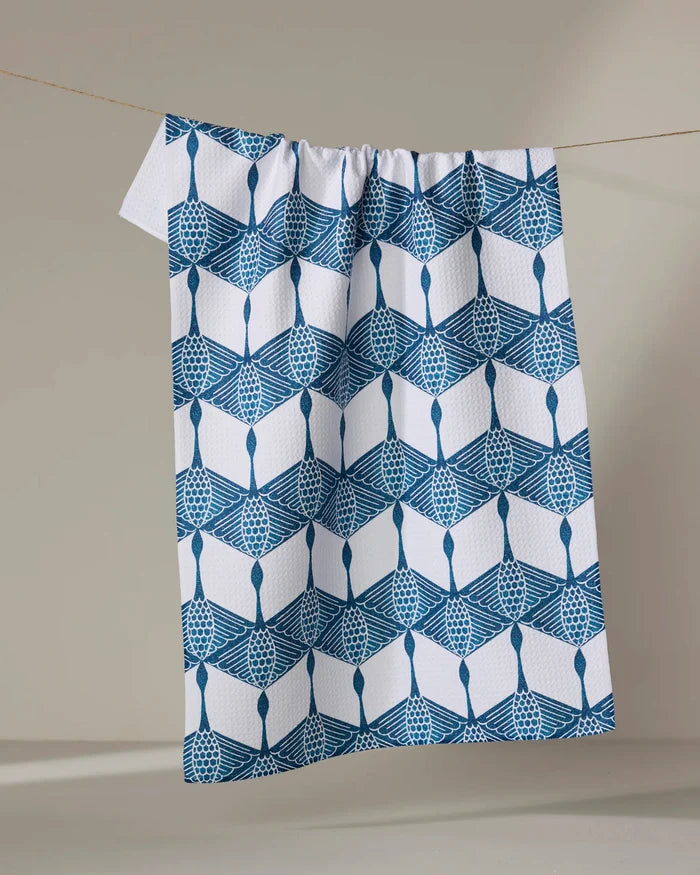 The BB Cubed kitchen towel by Renee Fly, made from recycled materials, features a blue geometric and lattice pattern on white and hangs from a clothesline against a neutral background.