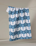 The BB Cubed kitchen towel by Renee Fly, made from recycled materials, features a blue geometric and lattice pattern on white and hangs from a clothesline against a neutral background.
