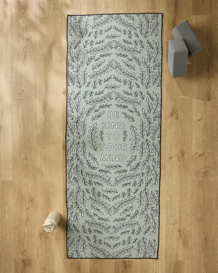 The Graphics and Grain "Be Kind To Your Mind" yoga mat with leafy patterns rests on a wooden floor, paired with a slip-resistant yoga towel, water bottle, and two stacked yoga blocks.