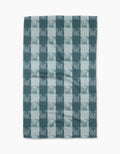 The Geometry “Beach Storm” blanket features a teal and light blue checkered pattern with subtle cherub accents on ultra-absorbent Geoweave™ fabric, creating a soft and cozy look ideal for relaxing moments.