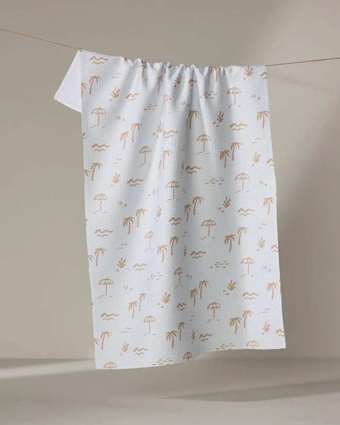 The "Beach" tea towel by Jen B. Peters is made from recycled materials and features a light brown beach pattern with palm trees, umbrellas, waves, and seagulls, shown hanging on a string against a neutral background.