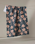The "Belgian Poppies" tea towel by Elisabeth Wing showcases a dark blue, quick-drying fabric with a peach botanical print of stylized, dandelion-like flowers, elegantly draped over a line against a neutral backdrop.