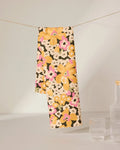 The GEOMETRY Beloved Bunch tea towel, featuring a vibrant floral design in yellow, pink, and white, hangs over a string near two clear glasses and part of a bottle on a plain surface.