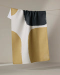 The Bend kitchen towel by Geometry features a textured abstract design in beige, white, and black, made from recycled materials, shown hanging on a clothesline against a neutral backdrop.