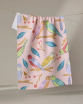 The "Berries and Budgies" tea towel by Elisabeth Wing, crafted from recycled materials, features colorful budgie birds, green leaves, and red berries on pink fabric, hanging on a string against a neutral background.