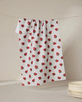 The Geometry Berry Patch tea towel, featuring a red strawberry pattern, hangs against a neutral backdrop beside stacked white bowls. Ultra-absorbent and stylish, it blends function with charm for any kitchen setting.