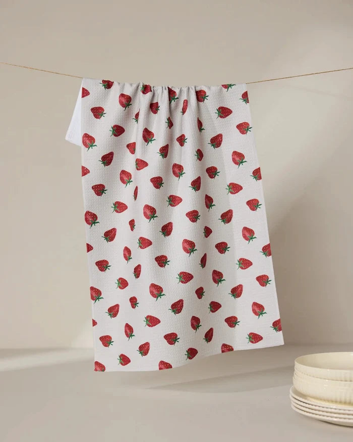 The Geometry Berry Patch tea towel, featuring a red strawberry pattern, hangs against a neutral backdrop beside stacked white bowls. Ultra-absorbent and stylish, it blends function with charm for any kitchen setting.