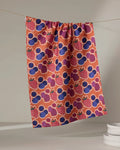 The Berrylicious tea towel by Lizzie Clark, made from recycled materials, features a quick-drying colorful strawberry and blueberry pattern. It hangs against a plain backdrop with white plates stacked in the lower right corner.
