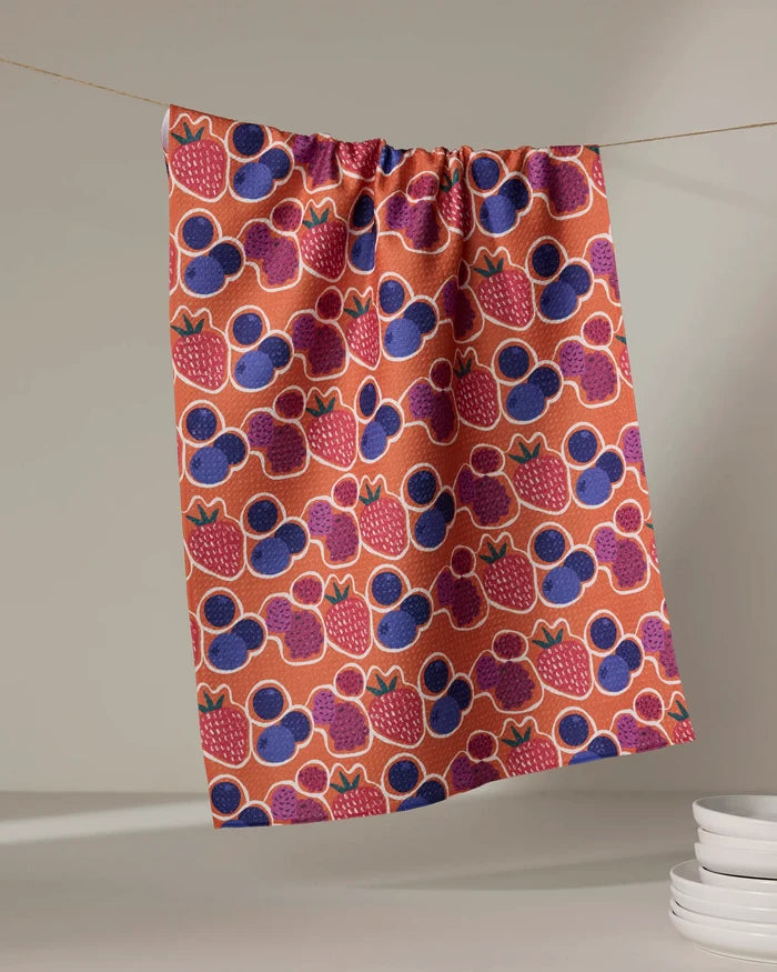 The Berrylicious tea towel by Lizzie Clark, made from recycled materials, features a quick-drying colorful strawberry and blueberry pattern. It hangs against a plain backdrop with white plates stacked in the lower right corner.