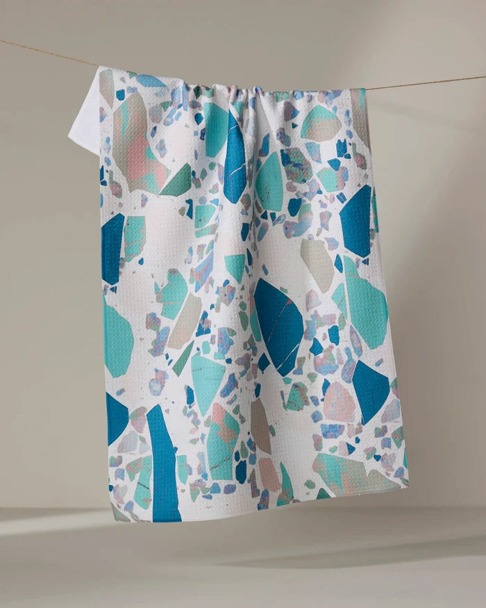 The Geometry Beryl tea towel, featuring a vibrant terrazzo pattern in blue, teal, and muted pastels, hangs on a clothesline against a neutral backdrop. Made from recycled materials, it's stylish and eco-friendly.