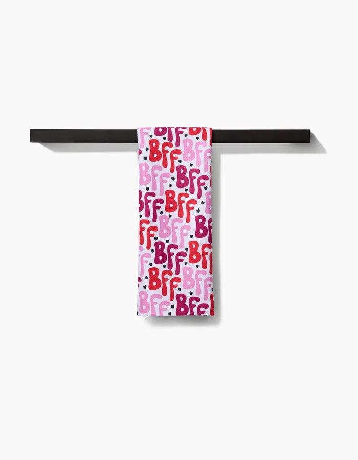A Hufton Studio Best Friends Forever tea towel hangs on a black bar, featuring a colorful BFF pattern in pink, red, and purple letters against a white background.