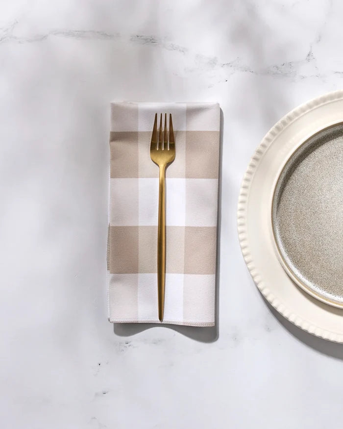 A gold fork rests on GEOMETRY’s eco-friendly "Betty Bakes" beige and white checkered napkin beside a decorative white plate on white marble.