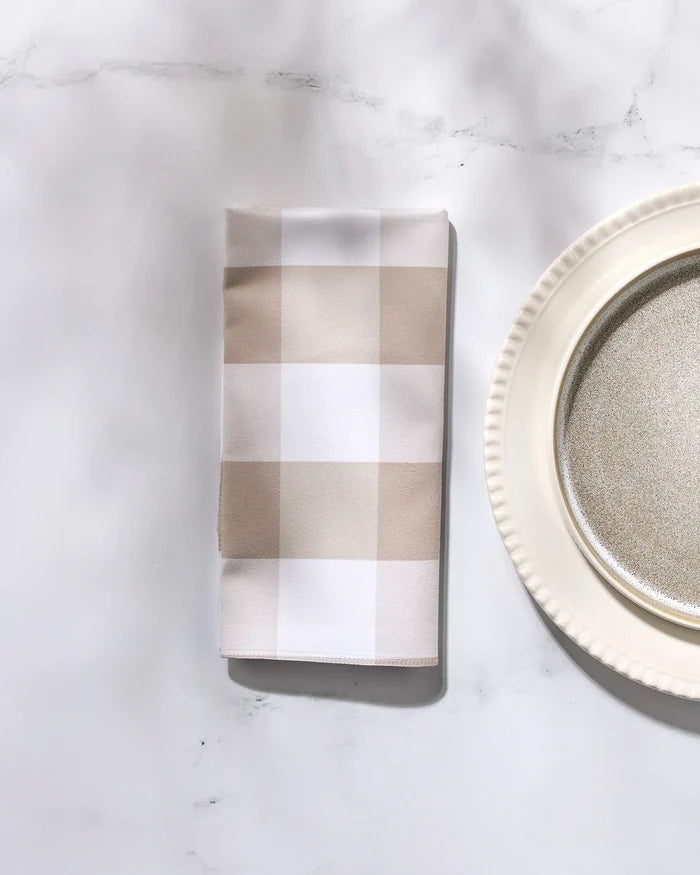 A folded GEOMETRY "Betty Bakes" beige and white checkered dinner napkin set rests on a white surface beside a round, cream-colored plate featuring a scalloped edge and metallic center.