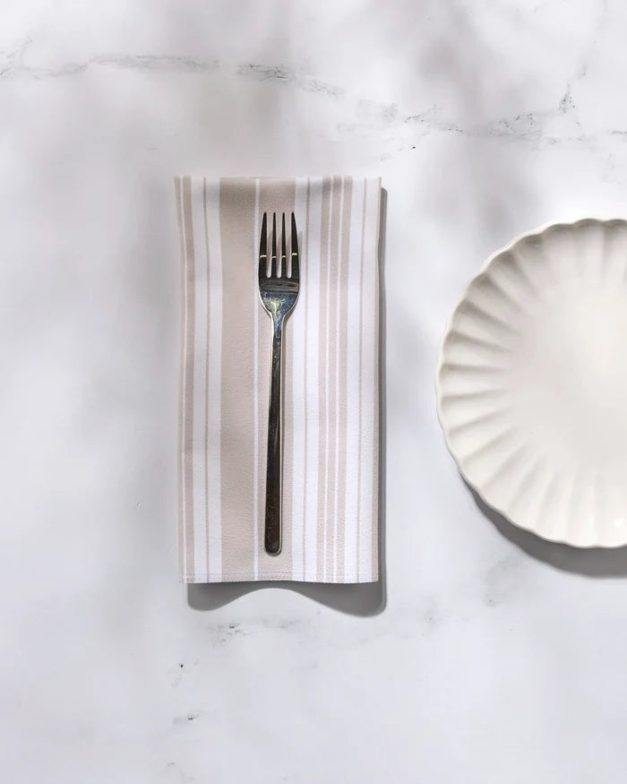 A GEOMETRY Betty Beach reusable dinner napkin is folded beneath a fork, set beside a white scalloped plate on a white marble surface.