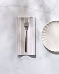 A GEOMETRY Betty Beach reusable dinner napkin is folded beneath a fork, set beside a white scalloped plate on a white marble surface.