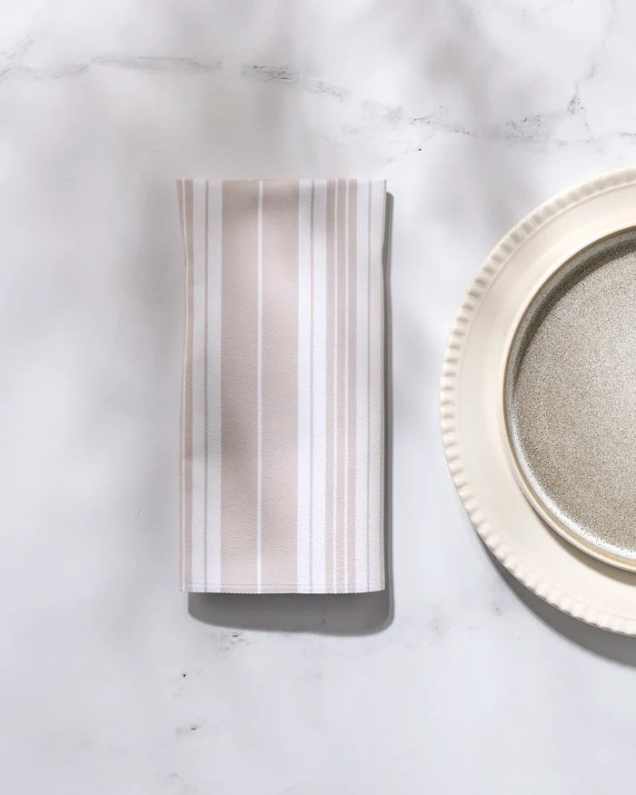 The GEOMETRY "Betty Beach" folded striped beige and white reusable dinner napkin is placed beside a cream plate with a metallic center on a white marble surface.