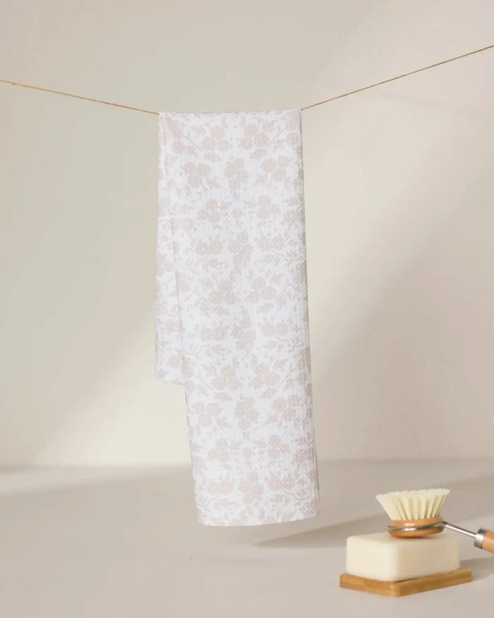 A light beige and white floral-patterned GEOMETRY tea towel, Bettys Buds, hangs on a string against a neutral background. In the foreground, an ultra-absorbent dish brush and a bar of soap rest on a dish.
