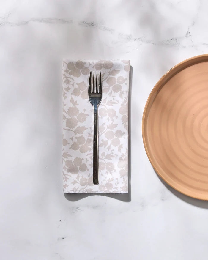 A metal fork rests on a folded GEOMETRY Betty's Buds floral-patterned recycled dinner napkin beside a round tan ceramic plate on a white marble surface with soft shadows.