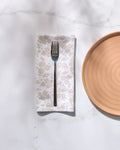 A metal fork rests on a folded GEOMETRY Betty's Buds floral-patterned recycled dinner napkin beside a round tan ceramic plate on a white marble surface with soft shadows.