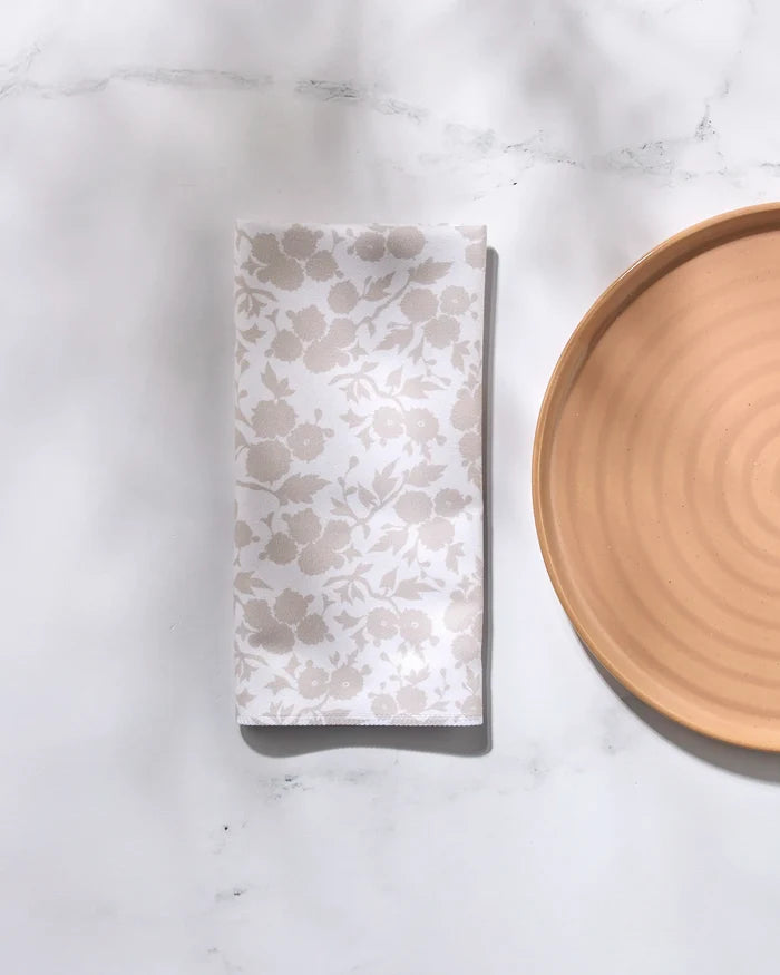 The GEOMETRY "Betty's Buds" beige ceramic plate with concentric circles sits on a white marble surface beside a folded recycled dinner napkin featuring a subtle floral pattern.