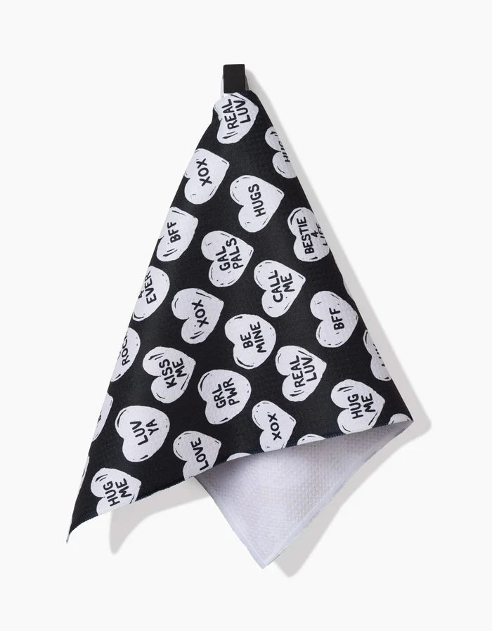 The BFF Candy Hearts bar towel by Hufton Studio features black ultra-absorbent fabric with white heart prints and phrases like REAL LUV, HUG ME, and XOX. It has a white backing and black hanging loop—charming for any space.