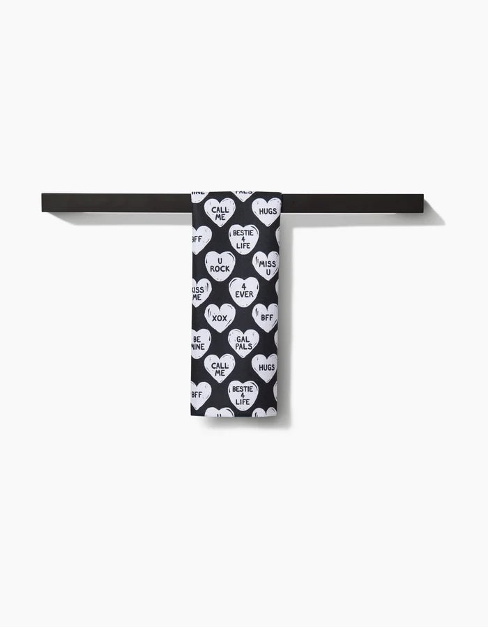 The Hufton Studio BFF Candy Hearts bar towel, black with white heart shapes and playful messages like BFF, HUGS, and CALL ME, hangs neatly on a dark bar against a plain white background.