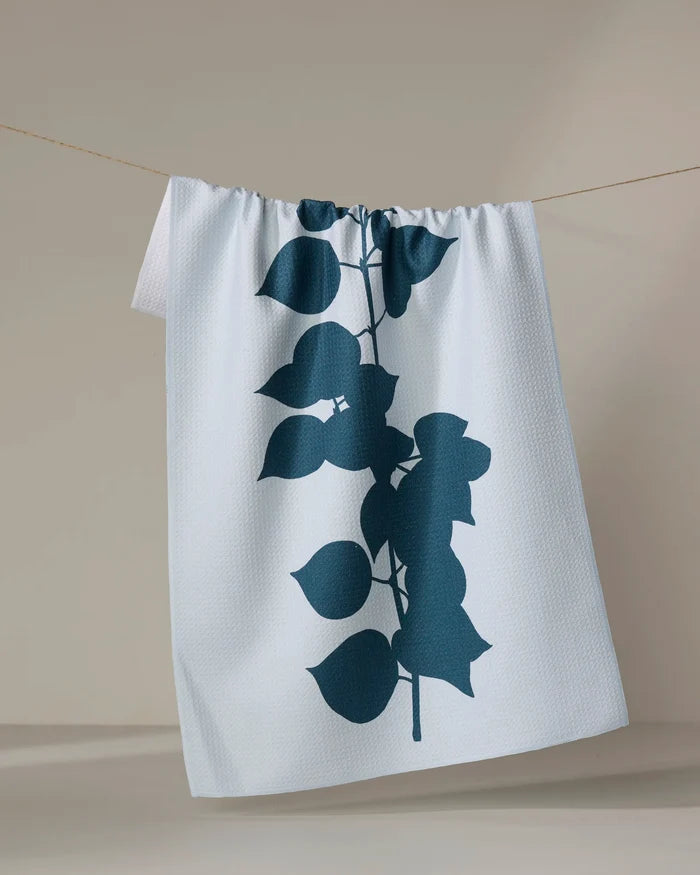The PrintedPlant Birch kitchen towel, featuring a white textured surface with a bold dark blue leafy branch design, hangs over a thin clothesline against a simple neutral backdrop.