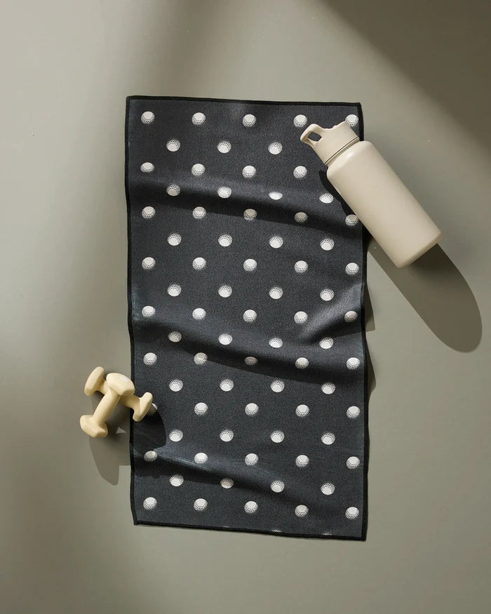 The Geometry Birdie fitness towel, black with white polka dots and made from recycled materials, lies flat on a light surface. A beige dumbbell and matching flip-top water bottle rest nearby the ultra-absorbent towel.