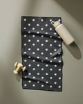 The Geometry Birdie fitness towel, black with white polka dots and made from recycled materials, lies flat on a light surface. A beige dumbbell and matching flip-top water bottle rest nearby the ultra-absorbent towel.